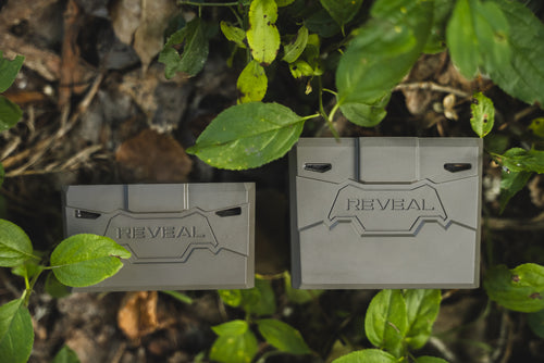 Two gray rectangular REVEAL branded boxes placed outdoors among green leaves and brown forest floor.