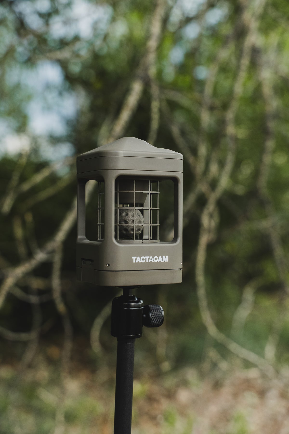 A wind direction indicator by TACTACAM mounted on a black pole, set against a blurred forest background.