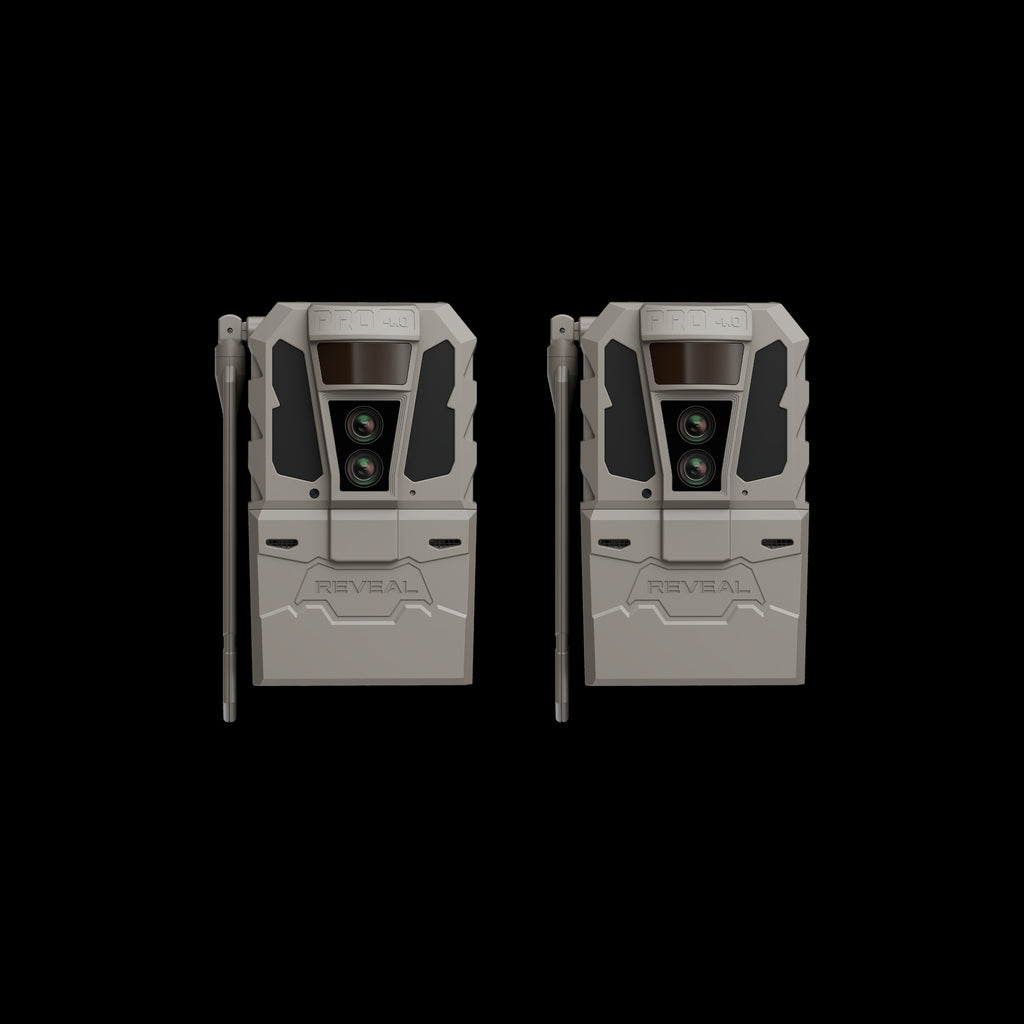 Two tactical trail cameras with a gray and black design, shown side-by-side against a solid black background. The cameras feature a front lens assembly, side antennas, and a lower compartment with the brand name 'REVEAL' embossed on the front.