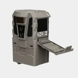 A tactical trail camera in a coyote brown color, featuring a hinged cover that is open to reveal a storage compartment. The camera has a rugged, angular design with dual lenses on the front and a screen on the side.